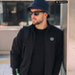 Mann mit Fischerhut und Sonnenbrille in schwarzer Zipper-Jacke mit Lorbeerkranz-Logo, Flasche in Tasche, khaki Cargohose.