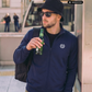 Mann mit Fischerhut und Sonnenbrille in blauer Zipper-Jacke mit Lorbeerkranz-Logo, hält eine Flasche, khaki Cargohose.