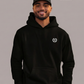 Mann mit Cap im schwarzen Hoodie mit Logo: Ein Lorbeerkranz umschließt ein kreisförmiges, gezacktes Symbol.