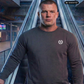 Kurzhaariger Mann im dunkelgrauen Sweatshirt mit Lorbeerkranz-Logo auf einer Rolltreppe im Bahnhofsbereich.