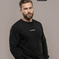 Bärtiger Mann im schwarzen Sweatshirt mit weißem Brustaufdruck „Standhaft“, Hände in den Taschen, grüne Hose.