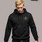 Ein Mann trägt einen schwarzen Kapuzenpullover mit einem kreisförmigen, gelb-schwarzen „STANDHAFT“-Logo samt kleiner Krone auf der linken Brust.