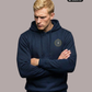 Ein Mann mit verschränkten Armen trägt einen dunkelblauen Kapuzenpullover mit einem kreisförmigen, gelb-blauen „STANDHAFT“-Logo samt kleiner Krone auf der linken Brust.