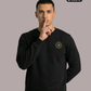 Ein Mann mit dem Zeigefinger vor dem Mund trägt einen schwarzen Pullover mit Rundhalsausschnitt und einem kreisförmigen, gelb-schwarzen „STANDHAFT“-Logo samt kleiner Krone auf der linken Brust.