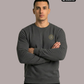 Ein Mann trägt einen dunkelgrauen Pullover mit Rundhalsausschnitt und einem kreisförmigen, gelb-grauen „STANDHAFT“-Logo samt kleiner Krone auf der linken Brust.