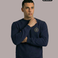 Ein Mann trägt einen dunkelblauen Pullover mit Rundhalsausschnitt und einem kreisförmigen, gelb-blauen „STANDHAFT“-Logo samt kleiner Krone auf der linken Brust.