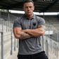 Ein Mann mit verschränkten Armen auf einer Stadiontribüne trägt ein graues Poloshirt mit einem schwarzen Wappen-Logo samt Adlergrafik und „STANDHAFT“-Schriftzug auf der linken Brust.