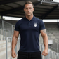 Ein Mann auf einer Stadiontribüne trägt ein dunkelblaues Poloshirt mit einem weißen Wappen-Logo samt Adlergrafik und „STANDHAFT“-Schriftzug auf der linken Brust.