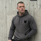 Ein Mann, der an einer Betonwand lehnt, trägt einen dunkelgrauen Hoodie mit einem schwarzen Wappen-Logo samt Adlergrafik und „STANDHAFT“-Schriftzug auf der linken Brust.