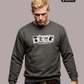Ein blonder Mann trägt einen grauen Pullover mit dem Aufruf „STANDHAFT“ und vier kleinen Karten Motiven sowie Jeans.