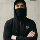 Mann mit Cap und Gesichtsmaske in schwarzer Zipper-Jacke mit Lorbeerkranz-Logo, verschränkte Arme, frontaler Blick.