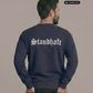 Ein Mann von hinten in einem dunkelblauen Pullover mit weißem „Standhaft“-Schriftzug im altdeutschen Stil auf dem Rücken.