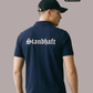 Ein Mann von hinten in einem dunkelblauen Poloshirt mit weißem „Standhaft“-Schriftzug im altdeutschen Stil auf dem Rücken.