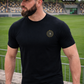 Ein Mann mit schwarzer Basecap im Stadion trägt ein schwarzes T-Shirt mit einem kreisförmigen, gelb-schwarzen „STANDHAFT“-Logo samt kleiner Krone auf der linken Brust.