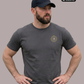 Ein Mann mit schwarzer Basecap und den Händen in den Hüften trägt ein dunkelgraues T-Shirt mit einem kreisförmigen, gelb-grauen „STANDHAFT“-Logo samt kleiner Krone auf der linken Brust.