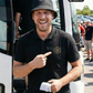 Ein lachender Mann mit schwarzem Anglerhut vor einem Bus trägt ein schwarzes Poloshirt mit einem kreisförmigen, gelb-schwarzen „STANDHAFT“-Logo samt kleiner Krone auf der linken Brust, auf das er mit dem Finger zeigt.