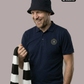 Ein Mann mit schwarzem Anglerhut und einem schwarz-weiß gestreiften Schal in der Hand trägt ein dunkelblaues Poloshirt mit einem kreisförmigen, gelb-blauen „STANDHAFT“-Logo samt kleiner Krone auf der linken Brust.