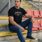 Ein Mann im Stadion trägt ein schwarzes T-Shirt mit goldenem „STANDHAFT“-Schriftzug und Lorbeerkranz-Logo auf der Brust.