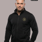 Ein Mann trägt eine schwarze Trainingsjacke mit Reißverschluss und einem kreisförmigen, gelb-schwarzen „STANDHAFT“-Logo samt kleiner Krone auf der linken Brust.