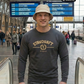 Ein Mann mit beigem Anglerhut trägt einen dunkelgrauen Pullover mit goldenem „STANDHAFT“-Schriftzug und Lorbeerkranz-Logo auf der Brust.