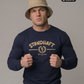 Ein Mann mit beigem Anglerhut trägt einen dunkelblauen Pullover mit goldenem „STANDHAFT“-Schriftzug und Lorbeerkranz-Logo auf der Brust.