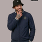 Ein Mann mit schwarzem Anglerhut trägt eine dunkelblaue Bomberjacke mit einem kreisförmigen, gelb-blauen „STANDHAFT“-Logo samt kleiner Krone auf der linken Brust.