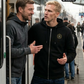 Zwei Männer stehen an einem Bahnsteig, wobei der rechte eine schwarze Zip-Jacke mit Kapuze und einem kreisförmigen, gelb-schwarzen „STANDHAFT“-Logo samt kleiner Krone auf der linken Brust trägt.
