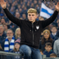 Ein Mann auf einem Stadionzaun mit erhobenen Händen trägt eine schwarze Zip-Jacke mit Kapuze und einem weißen Wappen-Logo samt Adlergrafik und „STANDHAFT“-Schriftzug auf der linken Brust.