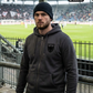 Ein Mann mit dunkler Strickmütze im Stadion trägt eine dunkelgraue Zip-Jacke mit Kapuze und einem schwarzen Wappen-Logo samt Adlergrafik und „STANDHAFT“-Schriftzug auf der linken Brust.