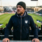 Ein bärtiger Mann mit dunkler Strickmütze im Fußballstadion trägt eine dunkelblaue Zip-Jacke mit Kapuze und einem weißen Wappen-Logo samt Adlergrafik und „STANDHAFT“-Schriftzug auf der linken Brust.